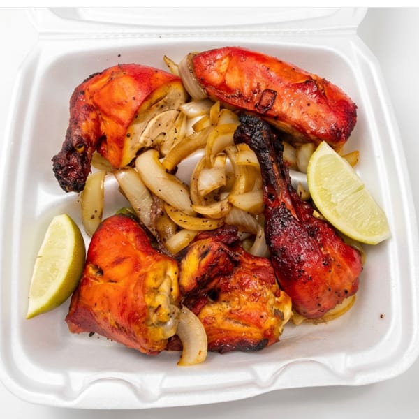 Tandoori chicken pieces with sautéed onions and lime wedges in a white takeout container.